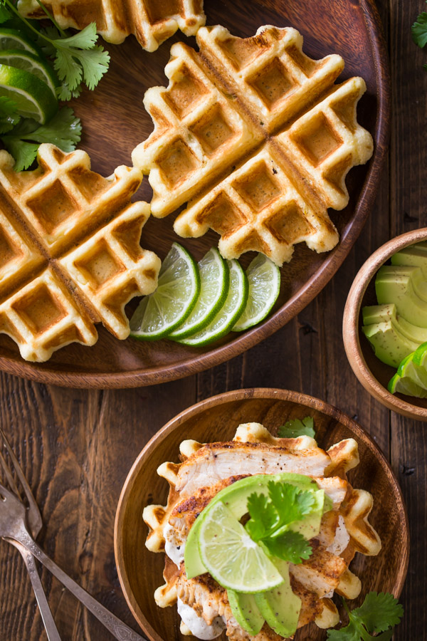 A Green Chile Chicken Waffletada assembled on a wood plate, sitting next to a large wood plate with mini cornbread waffles, lime slices and cilantro, and a small wood bowl of avocado slices.  