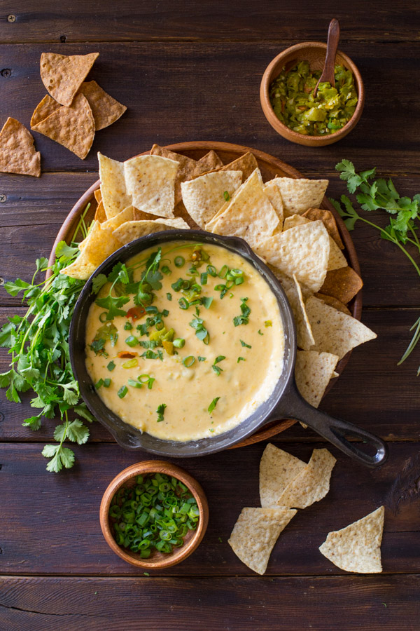 Hatch Green Chile Queso in a small cast iron skillet, garnished with green chiles, chopped cilantro, and green onions , sitting on a large wood plate with tortilla chips and a bundle of cilantro, with a small bowl of green onions and a small bowl of green chilies next to the plate.  