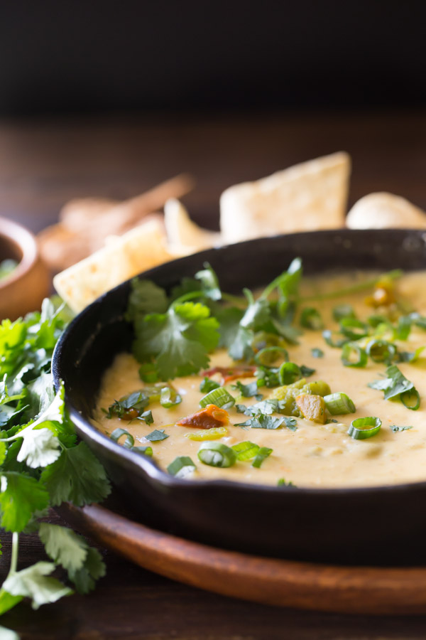 Hatch Green Chile Queso in a small cast iron skillet, garnished with green chiles, chopped cilantro, and green onions , sitting on a large wood plate with tortilla chips and a bundle of cilantro.