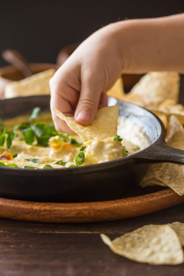 A tortilla chip being dipped into a cast iron skillet of Hatch Green Chile Queso that has been garnished with green chiles, chopped cilantro, and green onions.