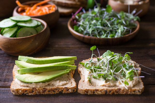 A Power Veggie Sandwich being assembled with roasted garlic hummus and slices of avocado on one slice of bread and roasted garlic hummus and micro greens on the other, with more ingredients for the sandwich in the background. 