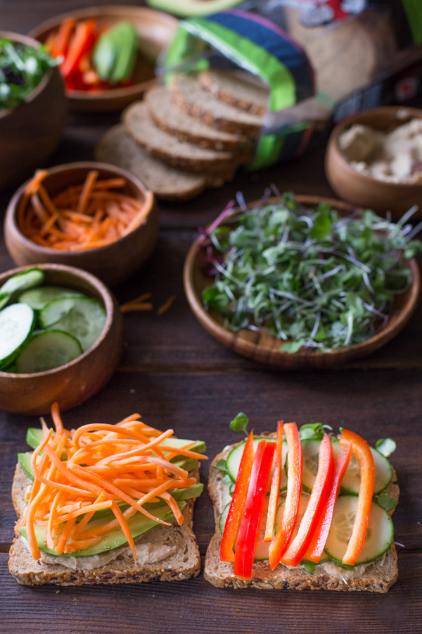 A Power Veggie Sandwich being assembled on two slices of bread, with all the ingredients in the background.  