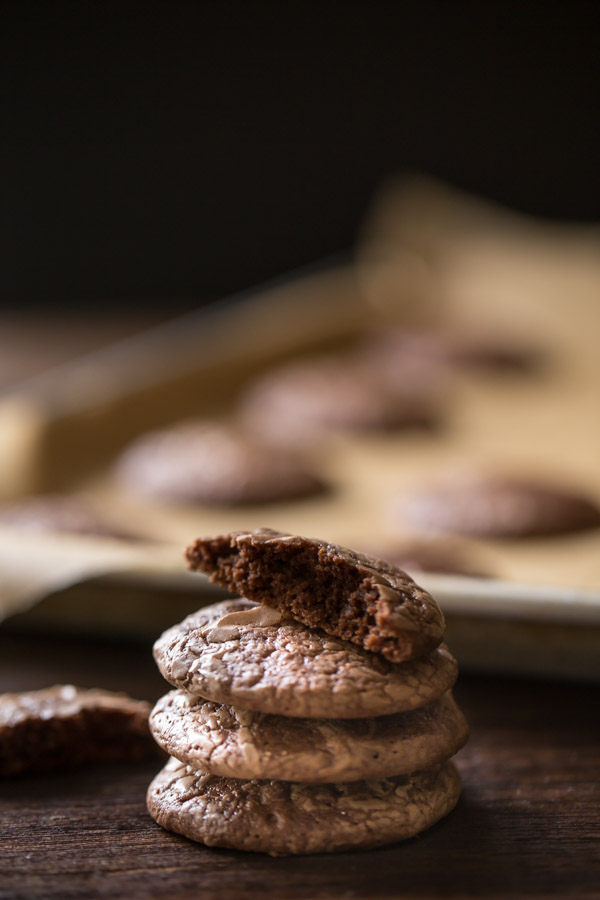 A stack of three Soft and Chewy Brownie Cookies with a half of a cookie on top, and more cookies on a parchment paper lined baking sheet in the background. 