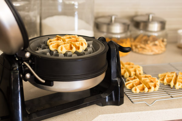 KitchenAid Waffle Baker with the lid open and a cooked mini cornbread waffle inside, with more waffles on a cooling rack next to it.  