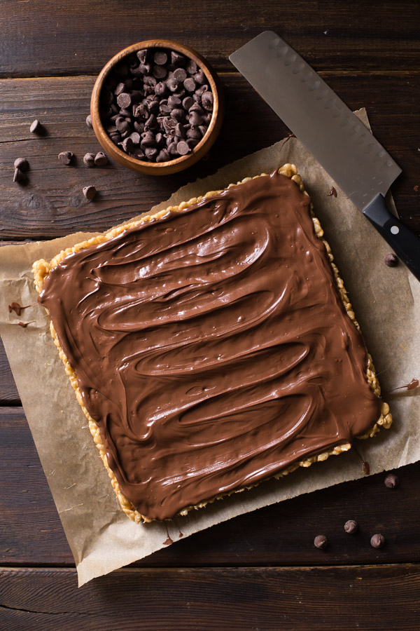 Chocolate Peanut Butter Rice Krispie Bars on a piece of parchment paper before being cut, with a small bowl of chocolate chips and a knife next to them.