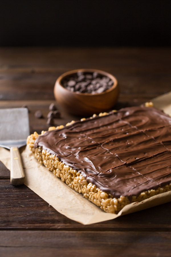 Chocolate Peanut Butter Rice Krispie Bars on a piece of parchment paper, with a small bowl of chocolate chips and a spatula next to them. 