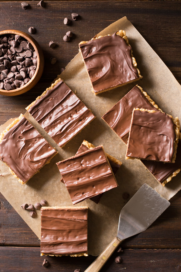 Chocolate Peanut Butter Rice Krispie Bars on a piece of parchment paper, with a small bowl of chocolate chips and a spatula next to them. 