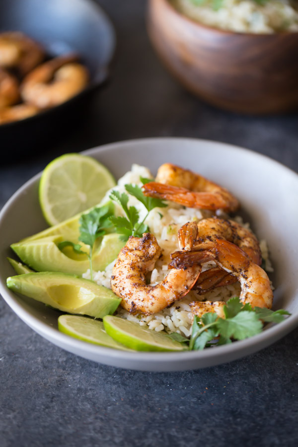 Cilantro Lime Rice Shrimp Bowl garnished with lime slices, avocado slices and fresh cilantro, with a wood bowl of Cilantro Lime Rice and a cast iron skillet of shrimp in the background.