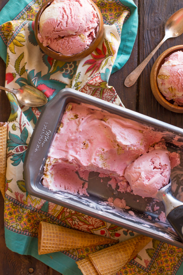 Homemade Raspberry Cheesecake Ice Cream in a loaf pan with an ice cream scoop, and two small wood bowls of Homemade Raspberry Cheesecake Ice Cream sitting next to it. 