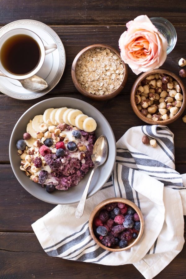 Triple Berry Oatmeal Breakfast Bowl topped with vanilla yogurt, crushed hazelnuts, sliced banana, and berries, with a cup of coffee, a bowl of oats, a bowl of hazelnuts and a bowl of berries next to the Triple Berry Oatmeal Breakfast Bowl.