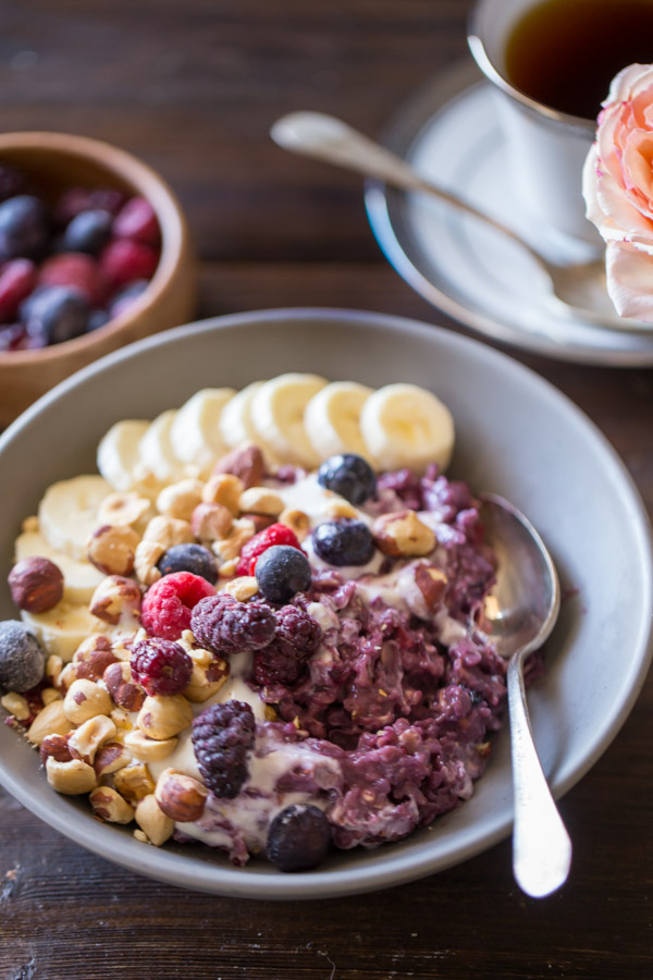 Triple Berry Oatmeal Breakfast Bowl topped with vanilla yogurt, crushed hazelnuts, sliced banana, and berries, with a small bowl of berries and a cup of coffee in the background.