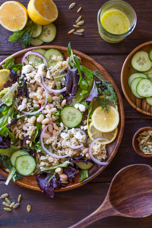 White Bean Couscous Salad With Lemon Vinaigrette on a large wood plate, with a wooden spoon, a small bowl of pepitas, a plate of sliced cucumbers, a glass jar of Lemon Vinaigrette, and a sliced lemon with some fresh dill and cucumber slices, all next to the plate of salad. 