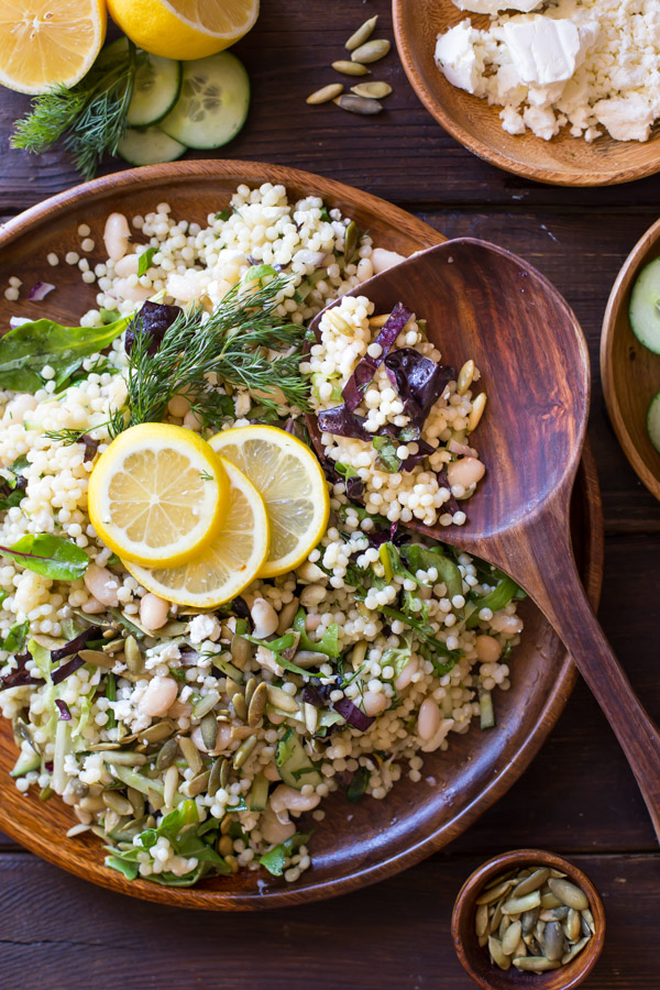White Bean Couscous Salad With Lemon Vinaigrette on a large wood plate, garnished with lemon slices and fresh dill, with a wooden spoon in it, and a wood bowl of feta cheese next to it along with a cut lemon with fresh dill and cucumber slices, and a small wood bowl of pepitas.  