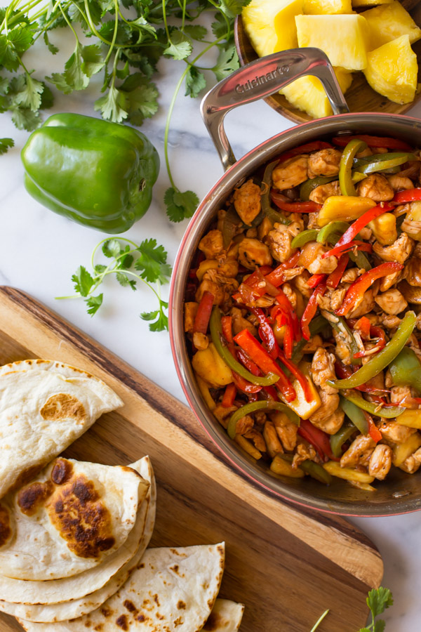 BBQ Chicken Fajitas in a large skillet, sitting next to a cutting board with browned tortillas, a whole green pepper, fresh cilantro and a plate of pineapple chunks. 