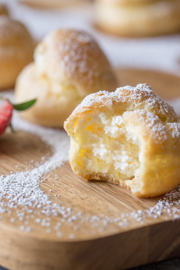 A Classic Cream Puff dusted with powdered sugar, with a bite taken out of it, sitting on a cutting board with more cream puffs.  