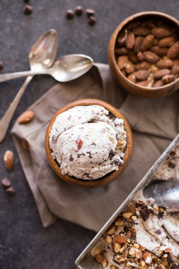 Dark Chocolate Almond Ice Cream in a small wood bowl, sitting next to two spoons, a small wood bowl of almonds and a loaf pan of Dark Chocolate Almond Ice Cream. 