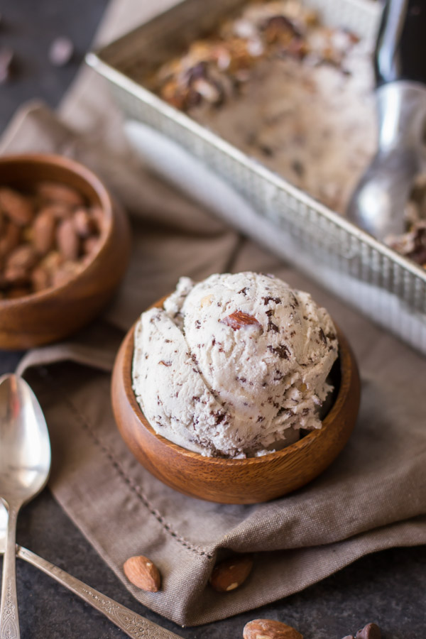 Dark Chocolate Almond Ice Cream in a small wood bowl, with a small wood bowl of almonds and a loaf pan of Dark Chocolate Almond Ice Cream in the background. 