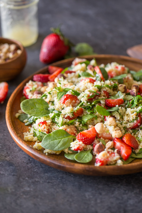 Strawberry Couscous Spinach Salad on a large wood plate, with a bowl of hazelnuts, a glass jar of almond vinaigrette and strawberries in the background.