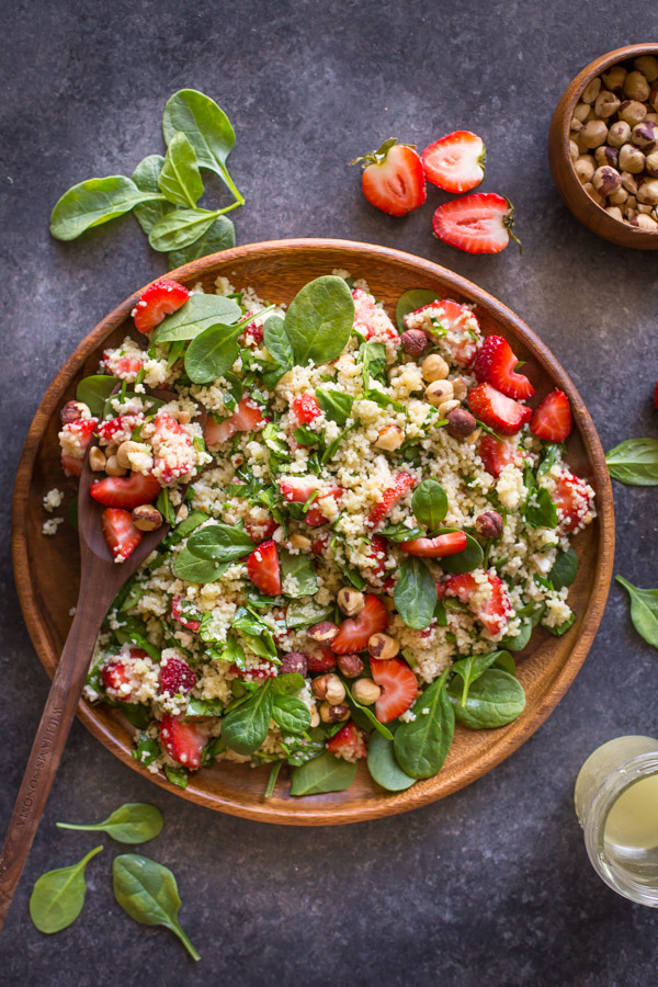 Strawberry Couscous Spinach Salad on a large wood plate with a wooden spoon it it, with spinach, strawberries, a bowl of hazelnuts and a glass jar of almond vinaigrette around the plate. 