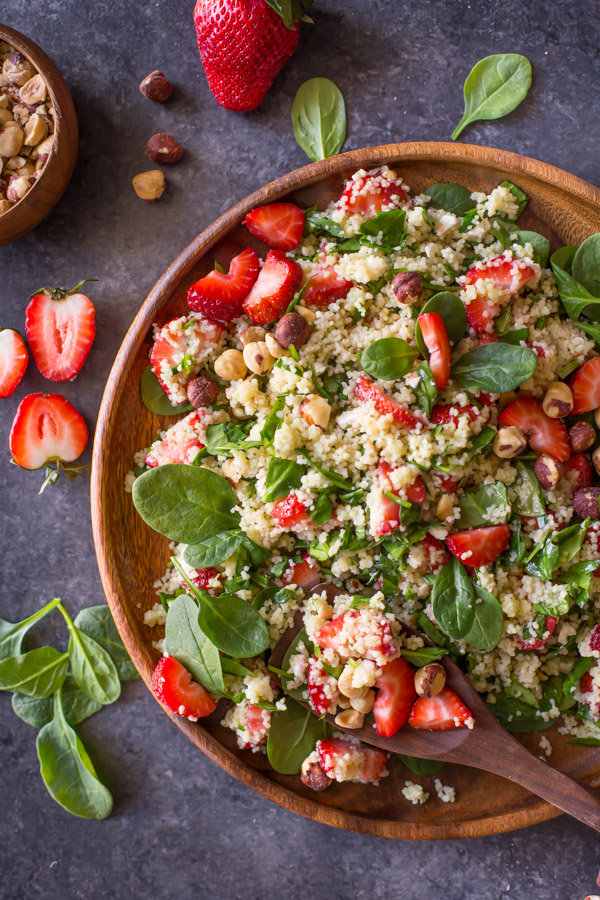 Strawberry Couscous Spinach Salad on a large wood plate with a wooden spoon it it, with spinach, strawberries and hazelnuts around the plate. 