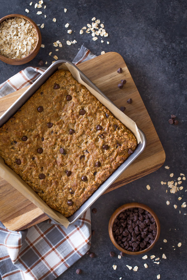Whole Wheat Oatmeal Chocolate Chip Snack Bars in a parchment paper lined baking pan, with a wood bowl of oats and a wood bowl of chocolate chips next to the baking pan. 