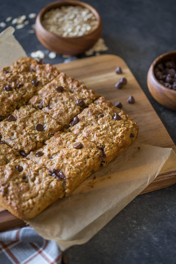 Whole Wheat Oatmeal Chocolate Chip Snack Bars on parchment paper sitting on a board, with a wood bowl of oats and a wood bowl of chocolate chips next to the board. 