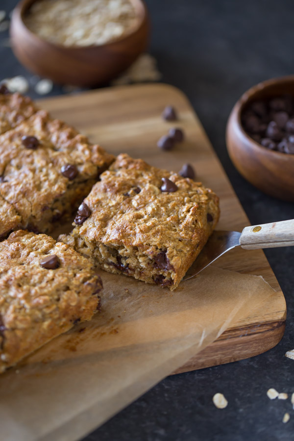 Whole Wheat Oatmeal Chocolate Chip Snack Bars on parchment paper sitting on a board, and a spatula under one of the bars, with a wood bowl of oats and a wood bowl of chocolate chips next to the board. 