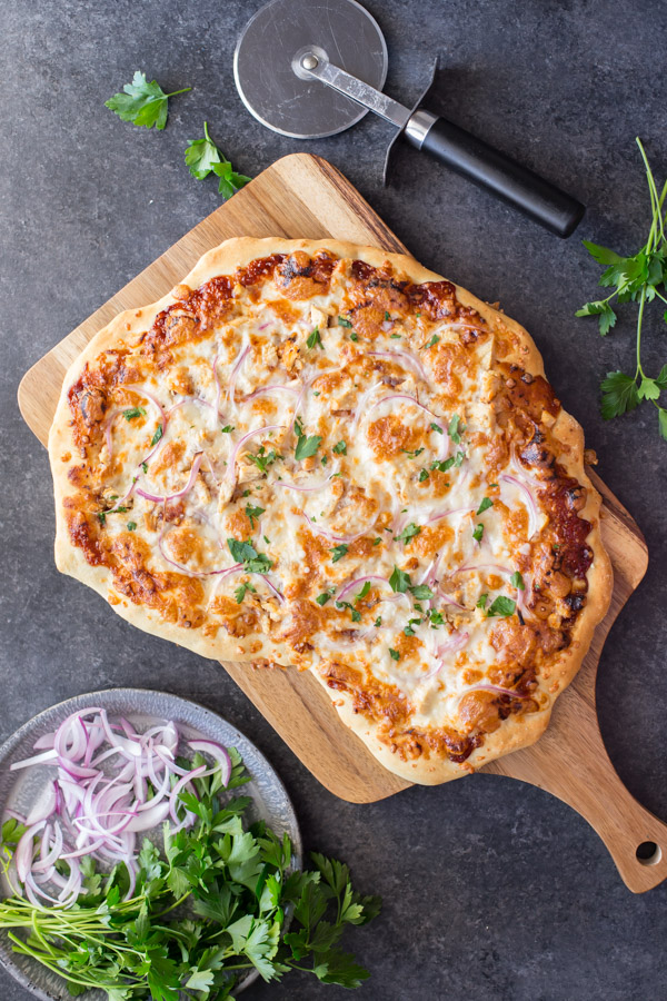 Barbeque Chicken Pizza on a cutting board, sitting next to a plate of flat leaf parsley and sliced red onions.  