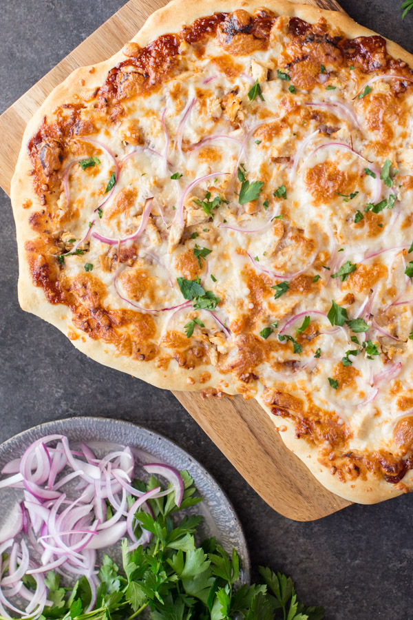 Barbeque Chicken Pizza on a cutting board, sitting next to a plate of flat leaf parsley and sliced red onions.  