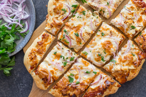 Barbeque Chicken Pizza sliced on a cutting board, sitting next to a plate of flat leaf parsley and sliced red onions.  