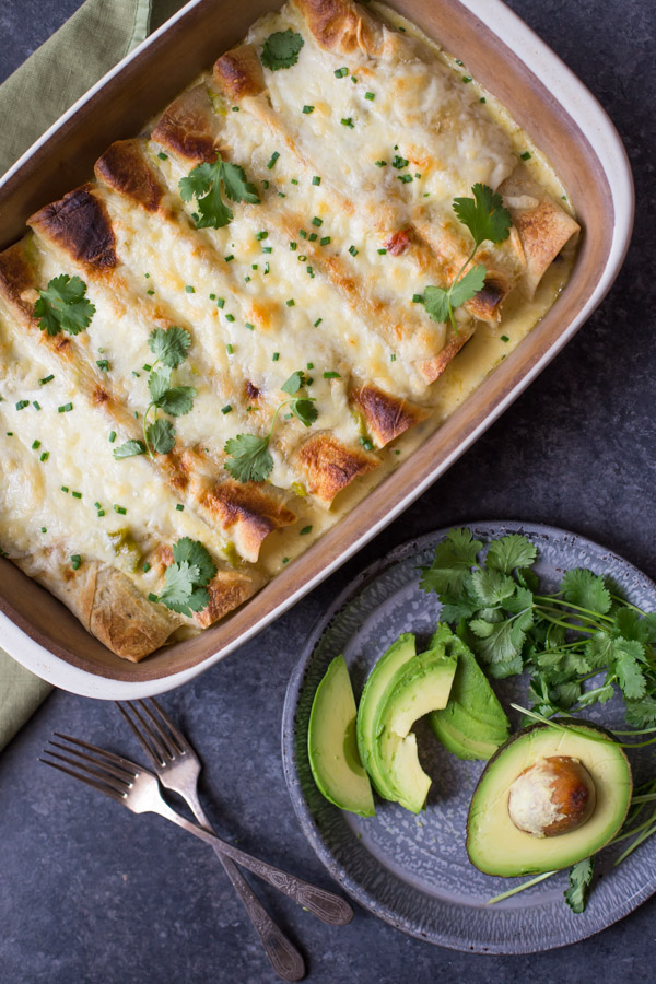 Hatch Green Chile Chicken Enchiladas in a baking dish garnished with cilantro, sitting next to a plate with avocado and cilantro, and two forks. 