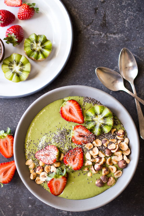 Strawberry Kiwi Protein Smoothie Bowl topped with sliced fresh strawberries and kiwi, hazelnuts, and chia seeds, with two spoons next to the bowl as well as a plate of strawberries and kiwi slices.