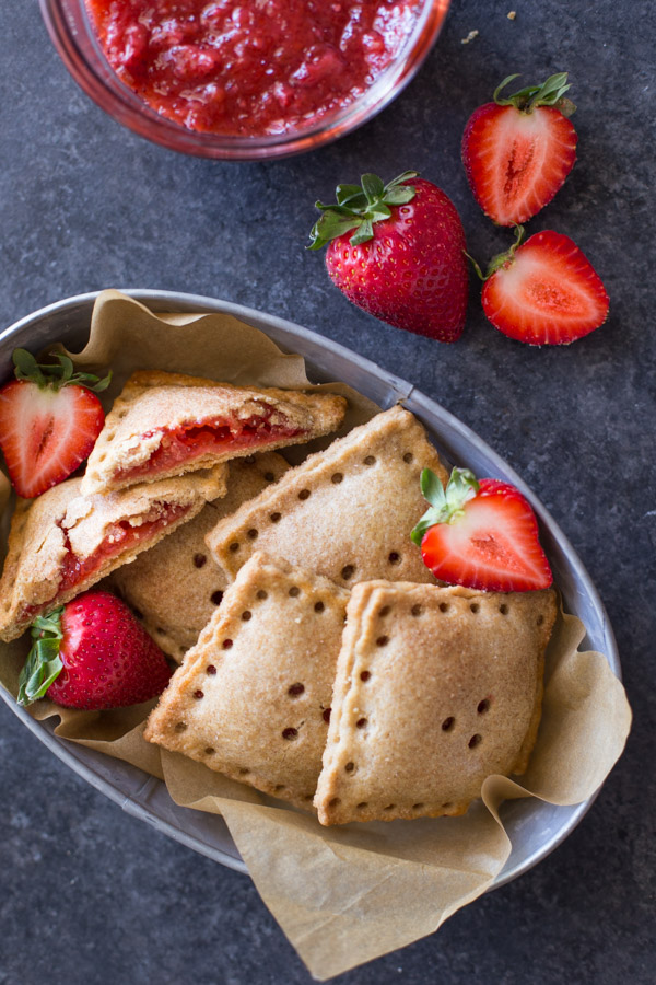 Whole Wheat Strawberry Rhubarb Fruit Pockets in a metal bowl, with some fresh strawberries in the bowl and next to it, along with a bowl of the strawberry rhubarb filling. 