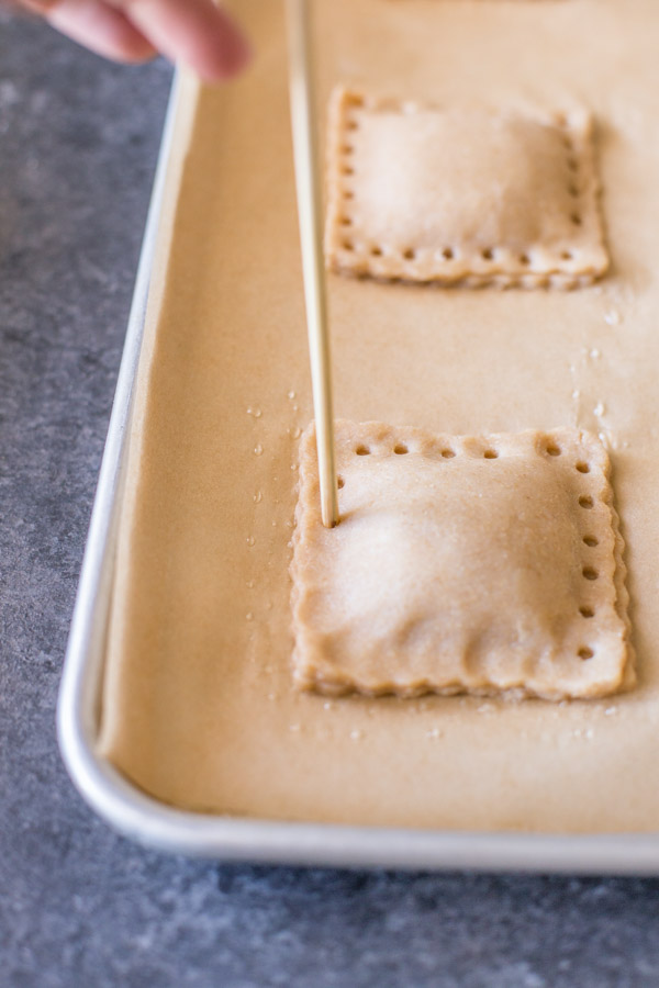 The blunt end of a wooden skewer being used to crimp the edges of the Whole Wheat Strawberry Rhubarb Fruit Pockets on a parchment paper lined baking sheet. 