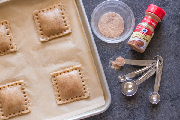 Whole Wheat Strawberry Rhubarb Fruit Pockets on a parchment paper lined baking sheet, with a bowl of cinnamon sugar, a spice container of McCormick Ground Cinnamon and some measuring spoons next to he baking sheet. 