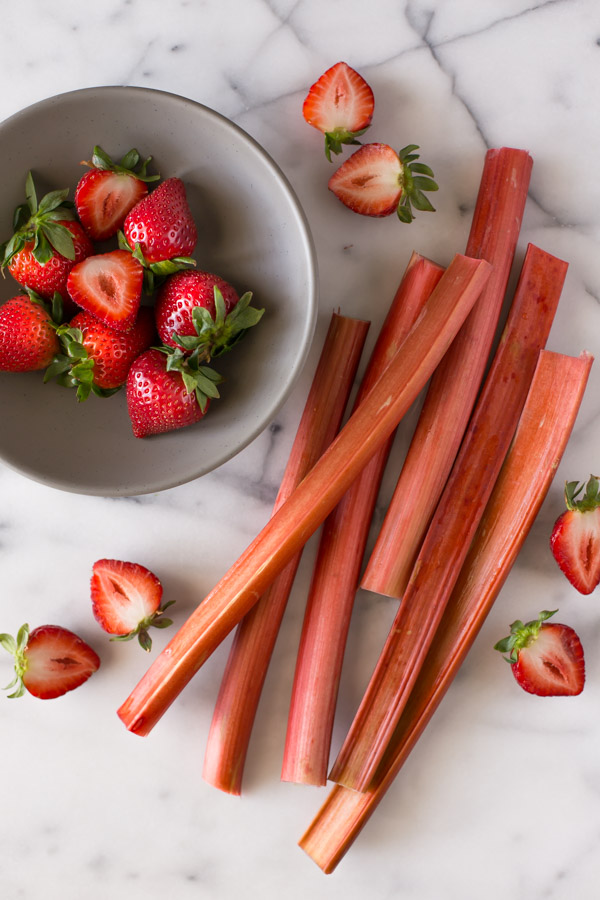 A bowl of strawberries sitting next to several rhubarb stalks and strawberries that have been cut in half.  