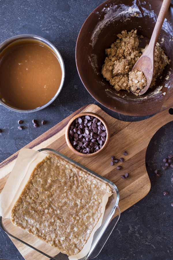 A parchment paper lined baking pan with the bottom layer of the oatmeal mixture in it, sitting on a cutting board with a small bowl of chocolate chips, with the sauce pan of caramel and the bowl of the remaining oatmeal mixture next to the cutting board. 