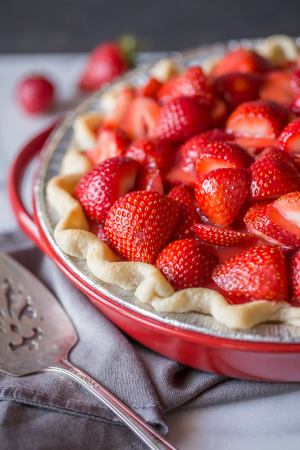 A Fresh Strawberry Pie in a pie plate, sitting on a cloth napkin with a serving utensil and whole strawberries next to it. 