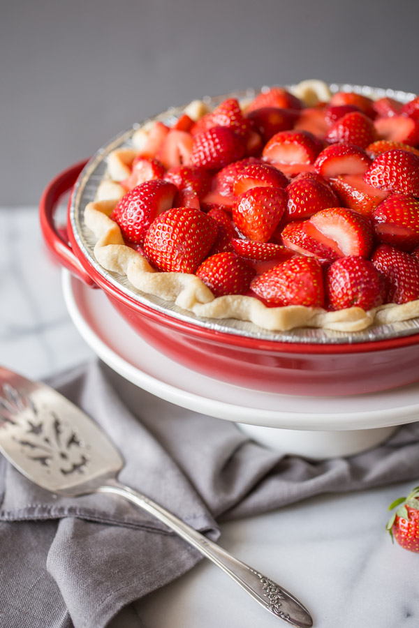 A Fresh Strawberry Pie in a pie plate, sitting on top of a cake stand, with a serving utensil next to it.  
