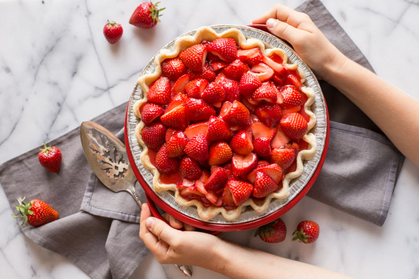 A Fresh Strawberry Pie in a pie plate, sitting on a cloth napkin with a serving utensil and whole strawberries next to it. 
