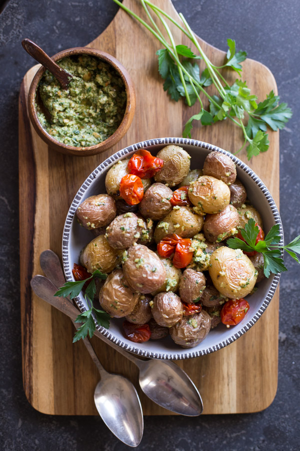 Roasted Baby Potatoes With Pesto in a serving bowl on a cutting board with a small wood bowl of Pesto, some fresh herbs and two spoons. 