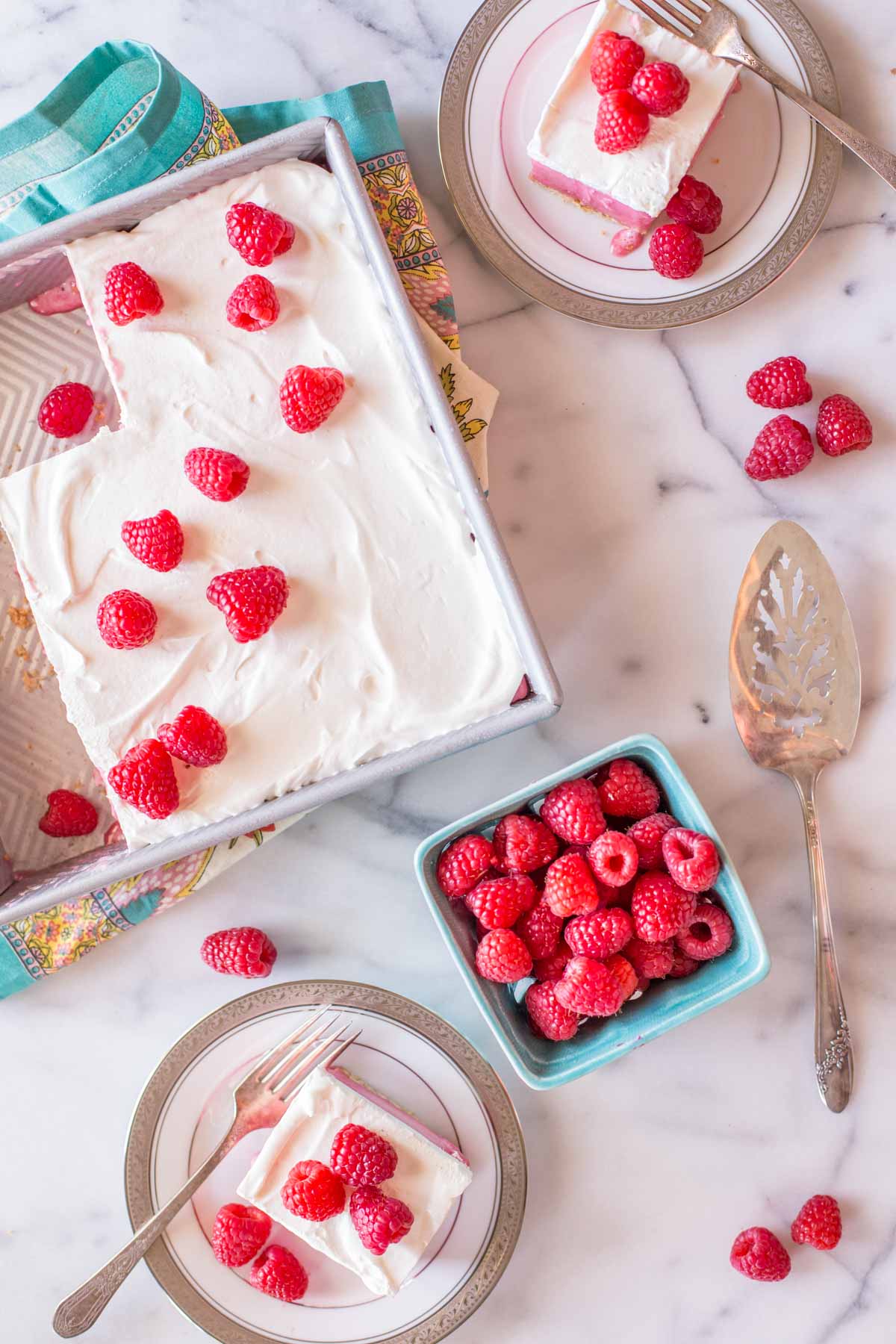 Cool and Creamy Raspberry Pretzel Dessert in a baking pan, sitting next to a carton of fresh raspberries and two plates with pieces of the dessert on each of them. 