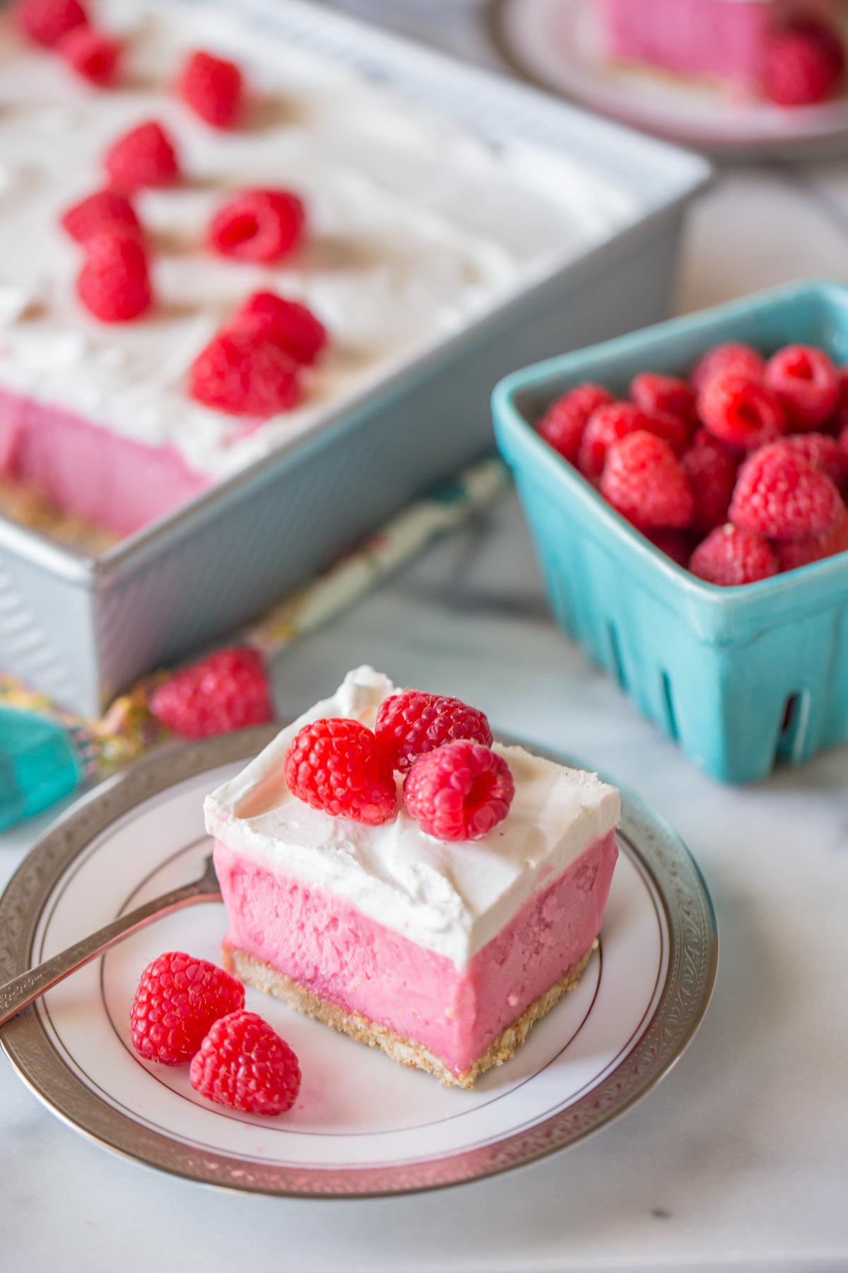 A piece of Cool and Creamy Raspberry Pretzel Dessert on a plate with fresh raspberries, and a carton of fresh raspberries in the background along with the baking pan of the Cool and Creamy Raspberry Pretzel Dessert. 
