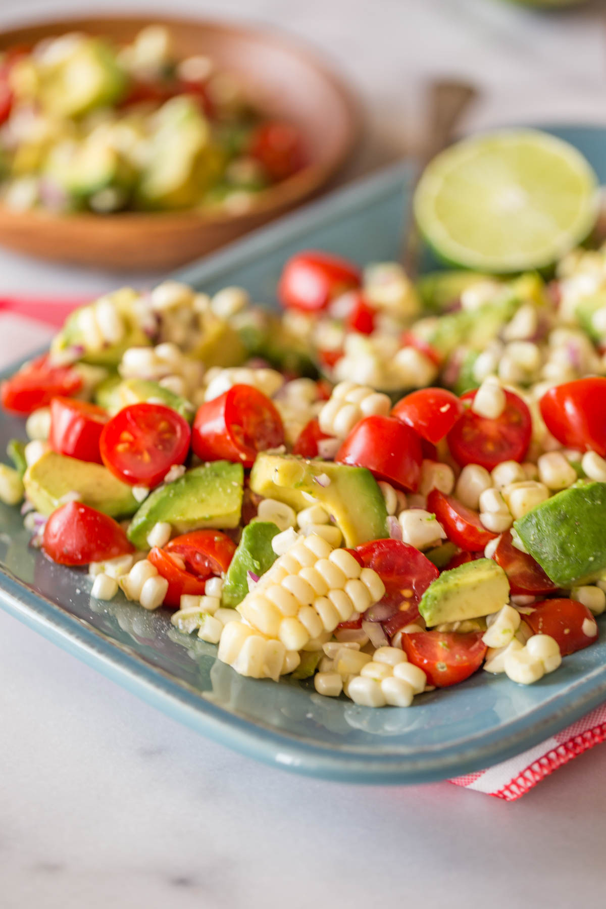 Corn, Tomato, and Avocado Salad on a platter, and a wood plate with a serving of the salad on it in the background. 