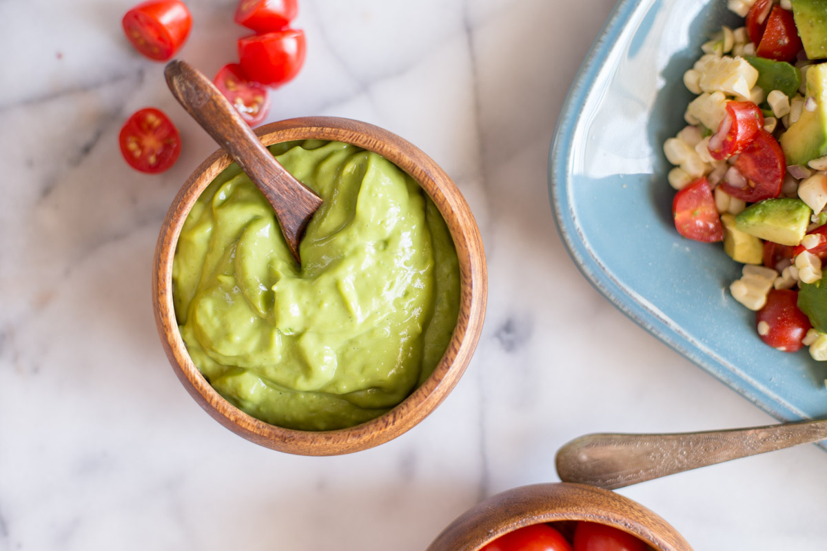 A small bowl of the avocado dressing with a little wooden spoon in it, sitting next to a platter of the Corn, Tomato, and Avocado Salad.