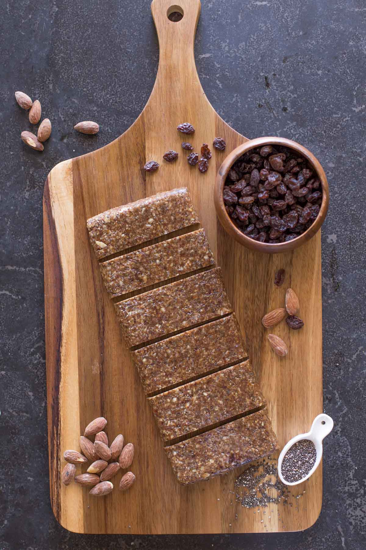 Five Ingredient No Bake Energy Bars on a cutting board, along with almonds, a small bowl of raisins and a spoon full of chia seeds. 