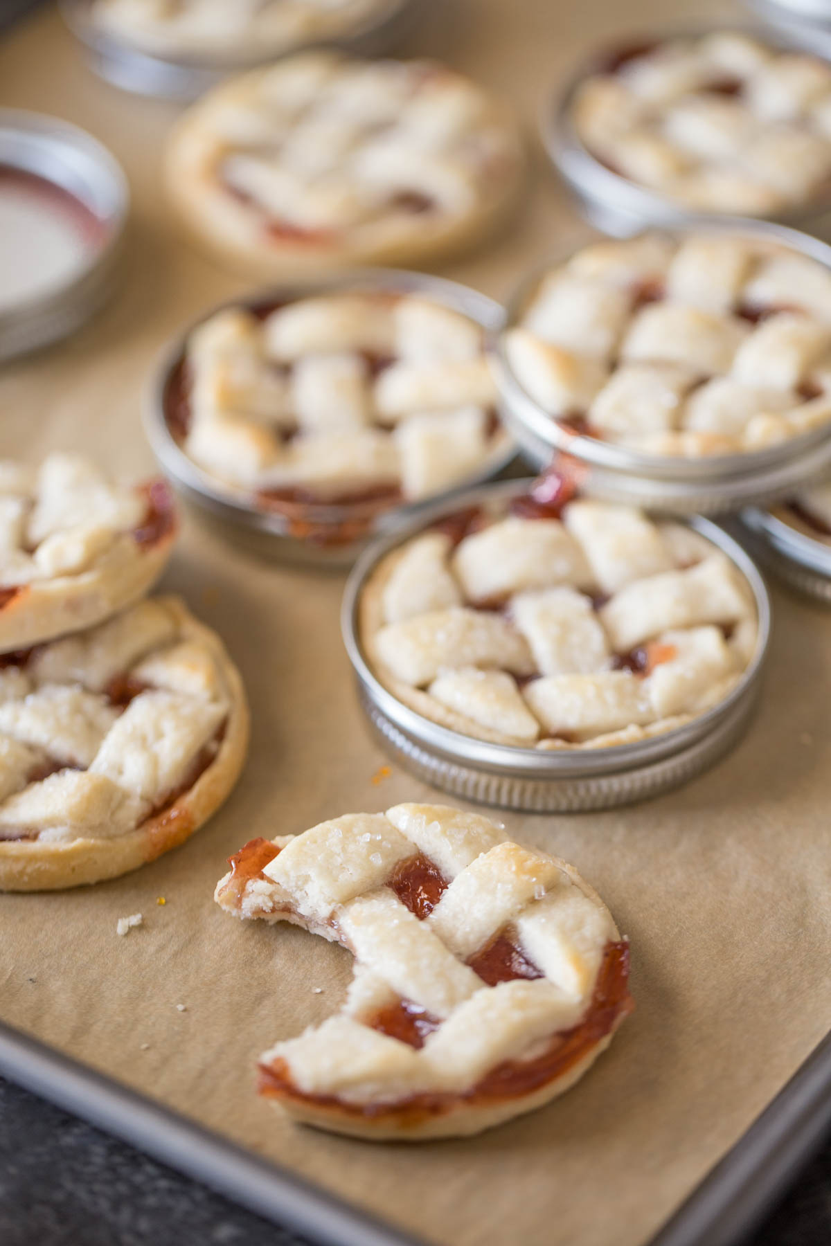 Mini Mason Jar Strawberry Pies on a parchment paper lined baking sheet after they have been baked, with some of the pies out of the lids and one pie with a bite taken out of it. 