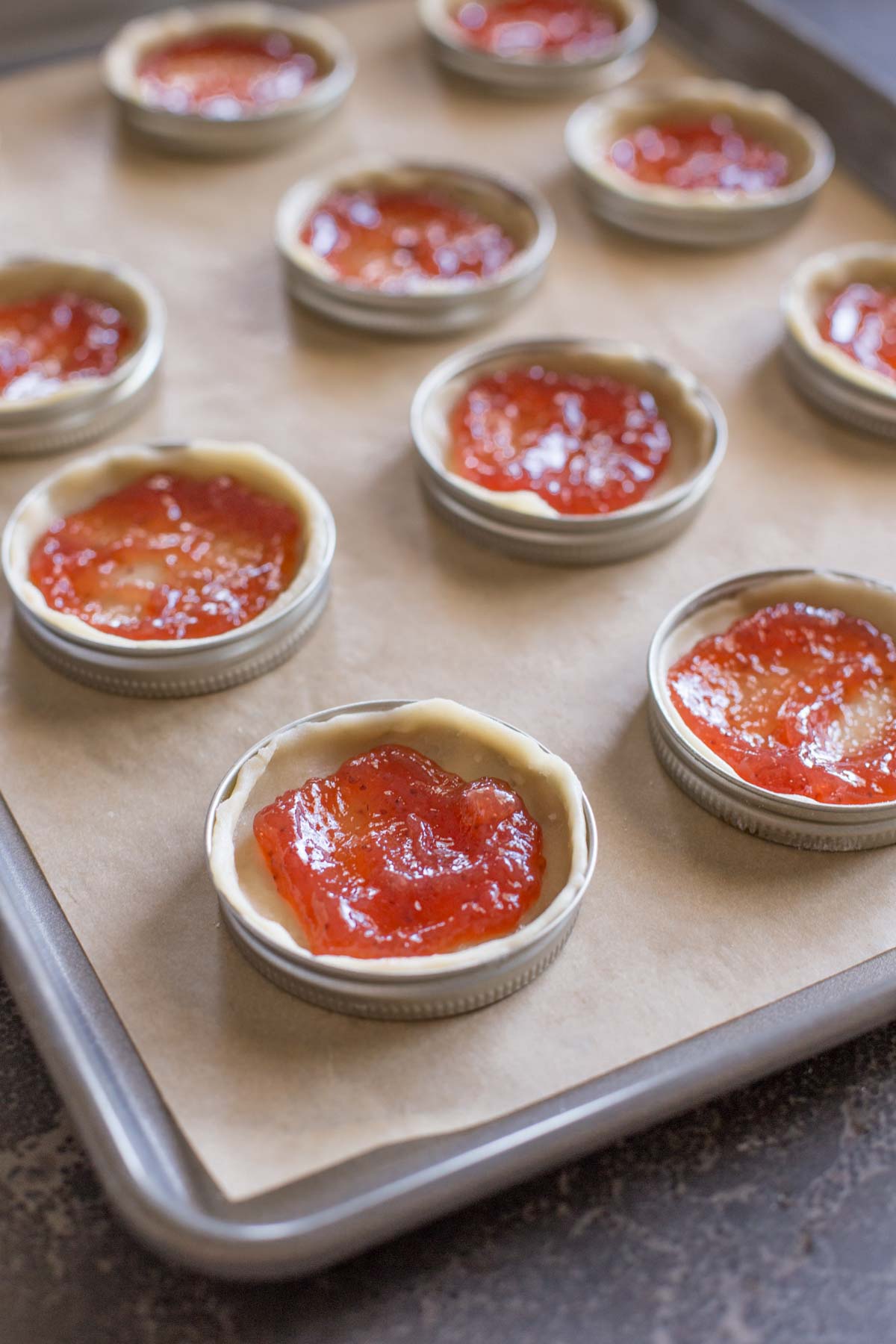 Mason jar lids arranged on a parchment paper lined baking sheet, with the pie dough and strawberry filling in them. 