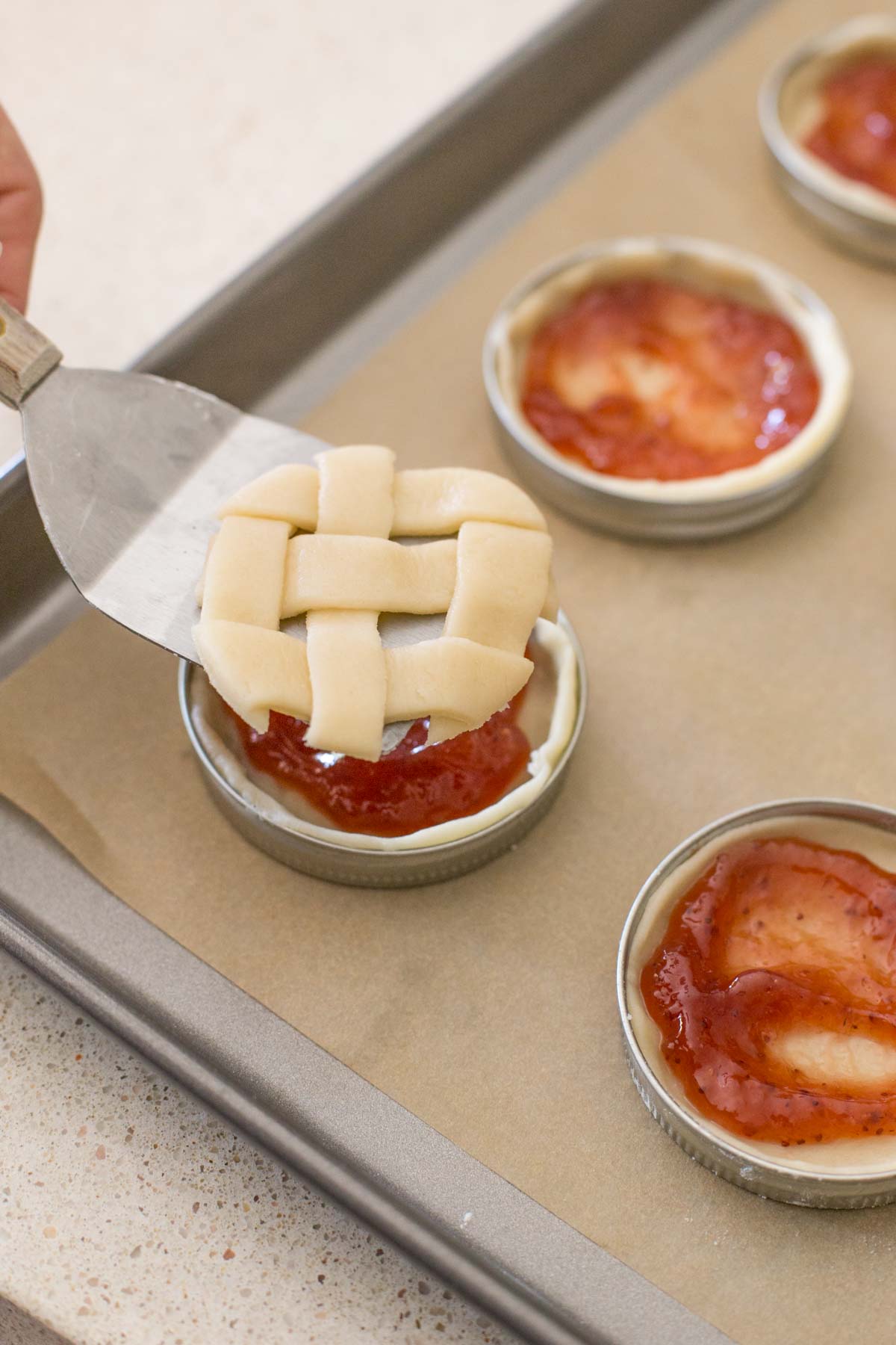 Mason jar lids arranged on a parchment paper lined baking sheet, with the pie dough and strawberry filling in them, and a metal spatula being used to transfer the circle lattice top onto the top of the pie.