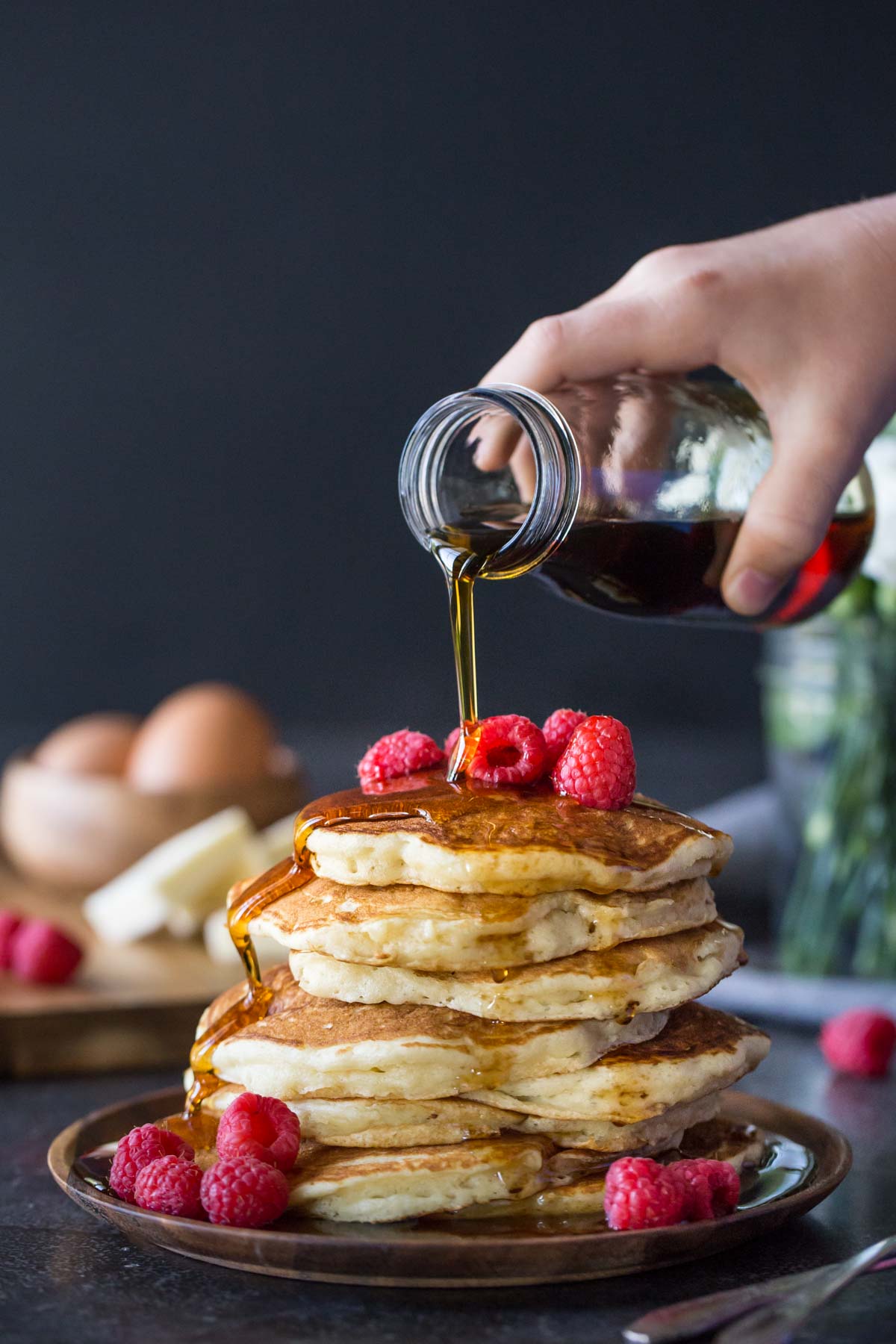 A stack of the Best Ever Buttermilk Pancakes on a plate, topped with fresh raspberries, with maple syrup being poured over them. 